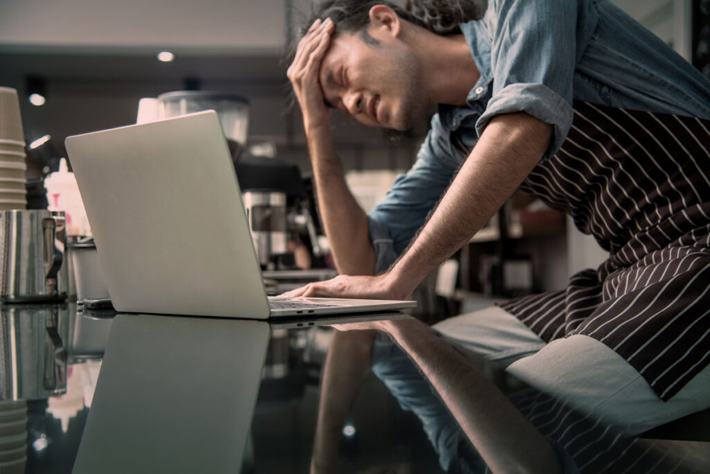 Barista frustrated at laptop while managing online orders in a busy coffee shop — metaphor for ecommerce stress